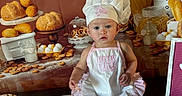 Amalia joined the competition — help win amazing prizes! apron, baby, baked_goods, bakery, baking, bread, chef_hat, child, cook, cute, dessert, eggs, food, indoors, kitchen, pastries, portrait, serious_expression, sitting, table