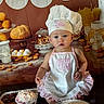 apron, baby, baked_goods, bakery, baking, bread, chef_hat, child, cook, cute, dessert, eggs, food, indoors, kitchen, pastries, portrait, serious_expression, sitting, table