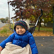 Benjamin is registered to the contest to win money with this photo: child, toddler, stroller, blue_jacket, knit_hat, blanket, autumn, fall_foliage, outdoor, smiling, happy, cute, cozy, park, seasonal, person, baby, warm_clothing, nature, daytime
