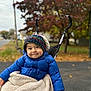 child, toddler, stroller, blue_jacket, knit_hat, blanket, autumn, fall_foliage, outdoor, smiling, happy, cute, cozy, park, seasonal, person, baby, warm_clothing, nature, daytime