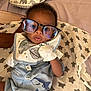 Harrison is registered to the contest to win money with this photo: baby, child, glasses, blue_outfit, bib, star_pattern, blanket, pillow, elephant_pattern, shark_patch, hand, face, curious, cute, indoor, laying, person, infant, soft_lighting, closeup