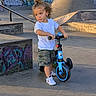 Bryson is registered to the contest to win money with this photo: toddler, child, balance_bike, skate_park, graffiti, concrete, outdoor, curly_hair, white_shirt, camouflage_shorts, sneakers, sunlight, shadow, ramp, fence, trees, person, playground, summer, urban