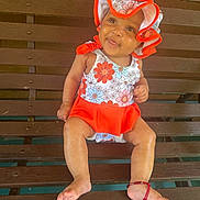 Alora is registered to the contest to win money with this photo: baby, child, bench, hat, floral_pattern, red_clothing, smile, barefoot, bracelet, wood, outdoor, cute, sitting, person, young_child, sunlight, happy, infant, fashion, portrait
