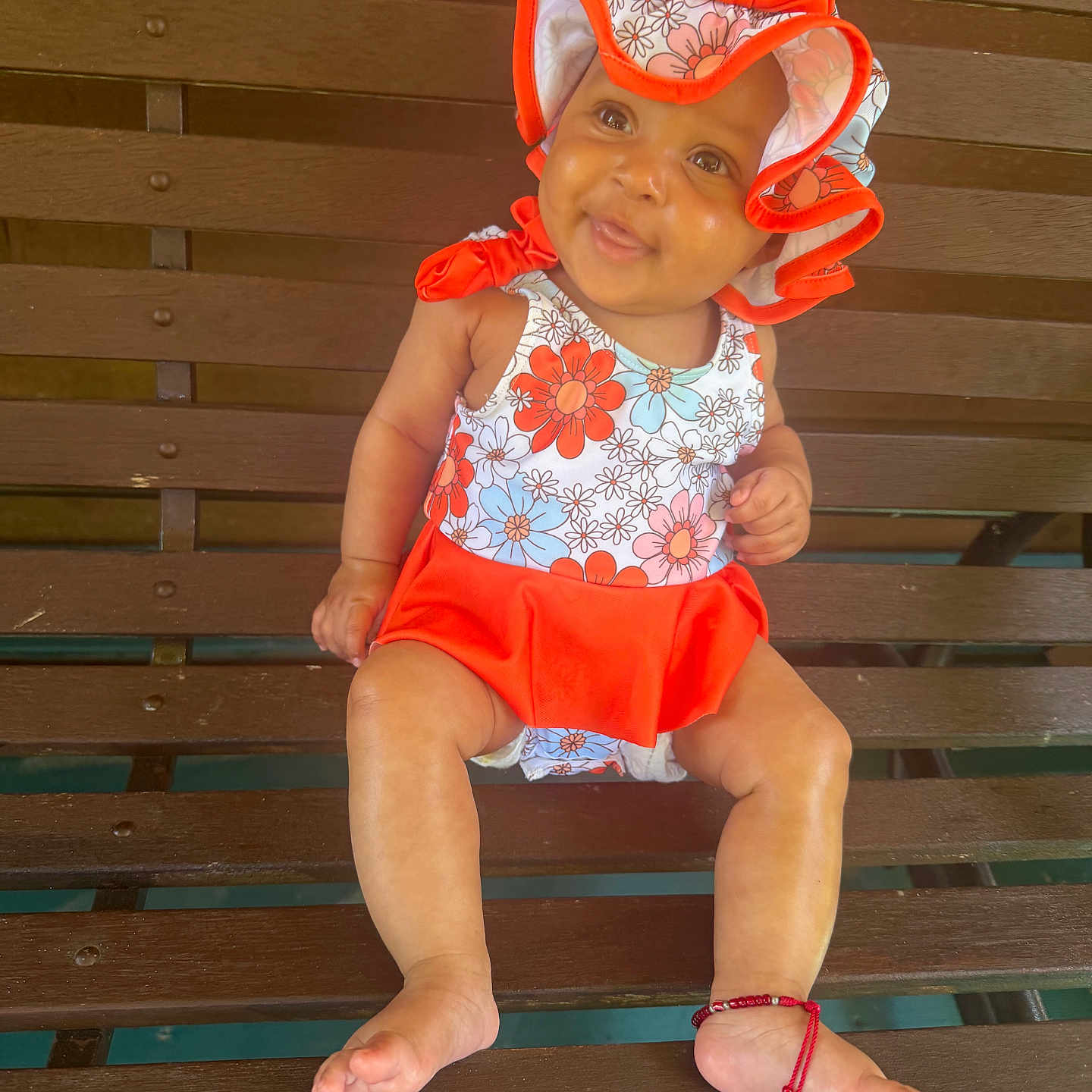 Alora is registered to the contest to win money with this photo: baby, child, bench, hat, floral_pattern, red_clothing, smile, barefoot, bracelet, wood, outdoor, cute, sitting, person, young_child, sunlight, happy, infant, fashion, portrait