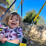 Piper is registered to the contest to win money with this photo: blurred_background, butterfly_pattern, child, clothing, daylight, face, fun, green_swing, hair, happy, metal_chain, nature, outdoor, playground, sky, smiling, swing, toddler, tree, yellow_swing