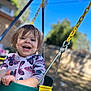 blurred_background, butterfly_pattern, child, clothing, daylight, face, fun, green_swing, hair, happy, metal_chain, nature, outdoor, playground, sky, smiling, swing, toddler, tree, yellow_swing