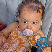 Wynter joined the competition — help win amazing prizes! baby, pacifier, curly_hair, orange_dress, floral_pattern, toy, unicorn_toy, teething_ring, child, infant, blue_teething_ring, cushion, seat, indoor, closeup, face, hand, expression, cute, childhood