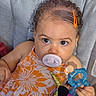 baby, pacifier, curly_hair, orange_dress, floral_pattern, toy, unicorn_toy, teething_ring, child, infant, blue_teething_ring, cushion, seat, indoor, closeup, face, hand, expression, cute, childhood