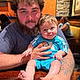 Beau is registered to the contest to win money with this photo: man, baby, child, beard, tattoo, blue_clothing, table, restaurant, wooden_table, smile, person, indoor, curly_hair, seated, holding, face, arm, casual_clothing, background, warm_lighting