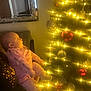 baby, child, christmas_tree, lights, ornaments, pink_clothing, calvin_klein, indoor, holiday, festive, decorations, sitting, soft_lighting, warm_glow, curious, face, person, cozy, reflection, mirror