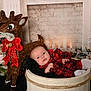 baby, infant, plaid_clothing, wooden_bucket, reindeer_decoration, red_bow, holiday_decor, christmas, wrapped_presents, candles, festive, cozy, indoor, black_blanket, pine_branches, face, cute, celebration, winter, smiling