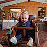 baby, child, high_chair, pink_headband, bows, smiling, navy_outfit, polka_dot, indoor, wooden_wall, living_room, toys, playpen, carpet, furniture, happy, person, footwear, home, family