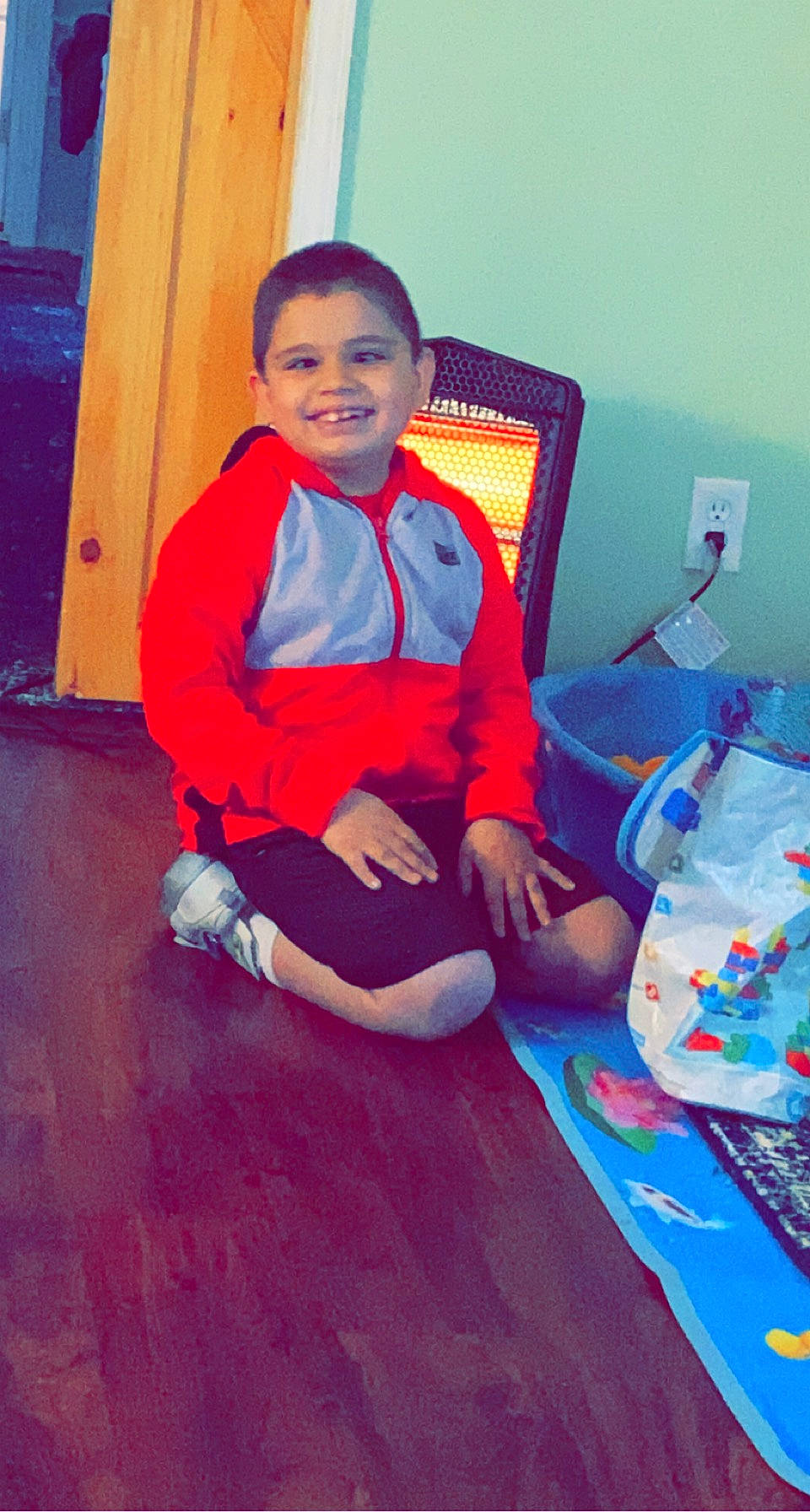 Cruz is registered to the contest to win money with this photo: comfort, door, elbow, electric_blue, face, floor, flooring, fun, happy, hardwood, human_leg, joy, knee, person, smile, sneakers, sock, t_shirt, thigh, toddler