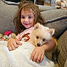 girl, child, puppy, dog, pet, cuddling, smiling, portrait, indoors, living_room, couch, blanket, plush_toy, fluffy, cream_colored, brown_hair, pink_shirt, sitting, cozy, family