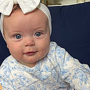 Ellie is registered to the contest to win money with this photo: baby, child, blue_eyes, bow, headband, floral_clothing, sitting, couch, indoor, portrait, cute, smile, hand, foot, face, infant, person, young_child, home_interior, soft_lighting