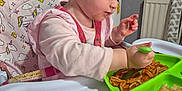 Eliza is registered to the contest to win money with this photo: toddler, child, high_chair, spaghetti, food, green_spoon, pink_clothing, unicorn_pattern, mealtime, messy, plate, feeding, indoor, baby, person, face, hair, hand, table, cereal