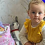 blonde_hair, blue_eyes, casual_clothing, child, grey_toy, home, indoor, long_sleeve, pink, plush_toy, portrait, serious_expression, standing, stuffed_animal, toddler, toy_bag, wallpaper, white_sleeves, yellow_dress, young_child
