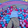 child, ball_pit, colorful_balls, pink_balls, blue_balls, red_balls, polka_dot_pants, white_shirt, love_text, bunny_graphic, outdoor, pavement, play, relaxation, happy_child, casual_clothing, toy, fun, kid, summer