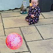 Eliza is registered to the contest to win money with this photo: toddler, child, pacifier, ball, outdoor, stone_patio, owl_statue, bin, curious, sitting, playful, daylight, casual_clothing, patterned_dress, barefoot, toy, cement, person, young_child, backyard
