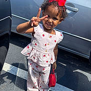 Knyla is registered to the contest to win money with this photo: toddler, child, smiling, peace_sign, red_bow, strawberry_pattern, outdoor, parking_lot, shoes, sandal, purse, sunlight, shadow, clothing, person, cute, happy, curly_hair, gesture, casual