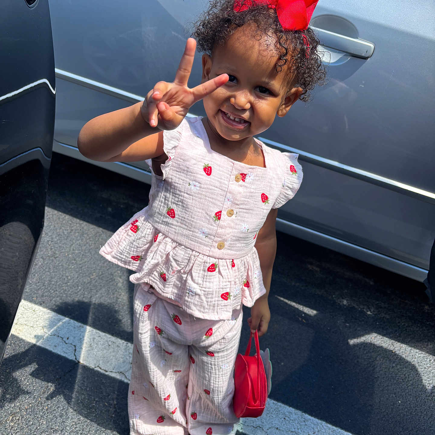 Knyla is registered to the contest to win money with this photo: toddler, child, smiling, peace_sign, red_bow, strawberry_pattern, outdoor, parking_lot, shoes, sandal, purse, sunlight, shadow, clothing, person, cute, happy, curly_hair, gesture, casual
