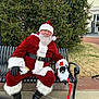santa_claus, dog, bench, christmas_tree, holiday, costume, outdoor, festive, beard, red_clothing, black_gloves, leash, grass, sidewalk, building, lights, hat, smile, person, pet