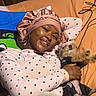 person, dog, small_dog, smiling, bedroom, bed, pajamas, bonnet, polka_dots, blanket, pillow, cuddling, pet, indoors, portrait, cozy, bedding, wood_paneling, charger_cable, tattoo