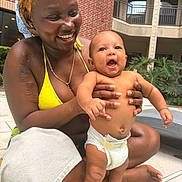 Ashton is registered to the contest to win money with this photo: woman, baby, smiling, happy, outdoor, poolside, diaper, holding, person, bikini, skin, joy, toddler, building, brick_wall, greenery, sunlight, nails, tattoo, accessory