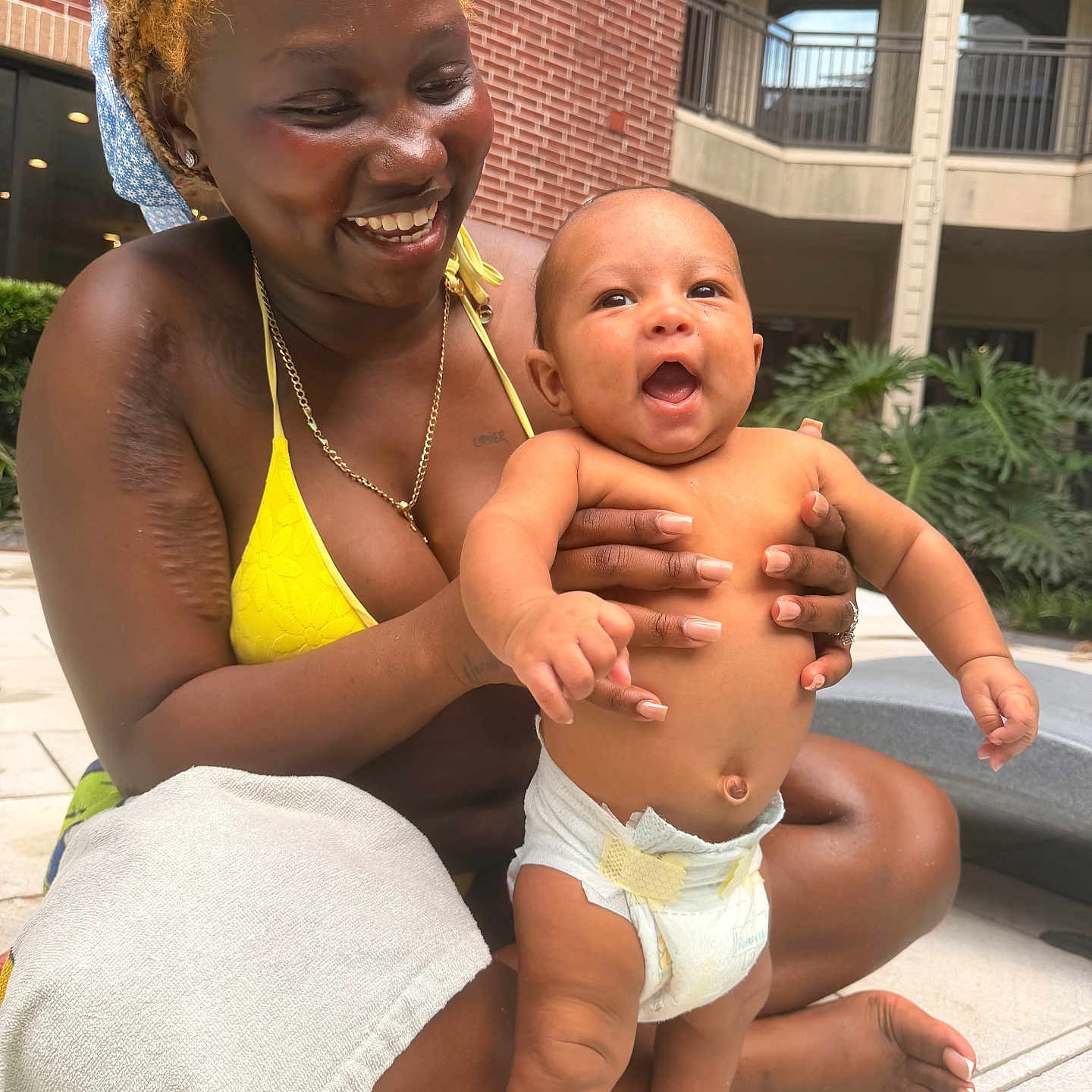 Ashton is registered to the contest to win money with this photo: woman, baby, smiling, happy, outdoor, poolside, diaper, holding, person, bikini, skin, joy, toddler, building, brick_wall, greenery, sunlight, nails, tattoo, accessory