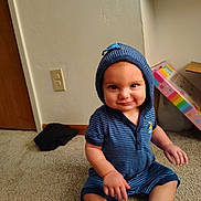 Luther joined the competition — help win amazing prizes! baby, child, toddler, sitting, smiling, happy, portrait, face, blue_outfit, striped_clothing, hat, hood, carpet, indoor, home, door, wall, electrical_outlet, colorful_package, bare_feet
