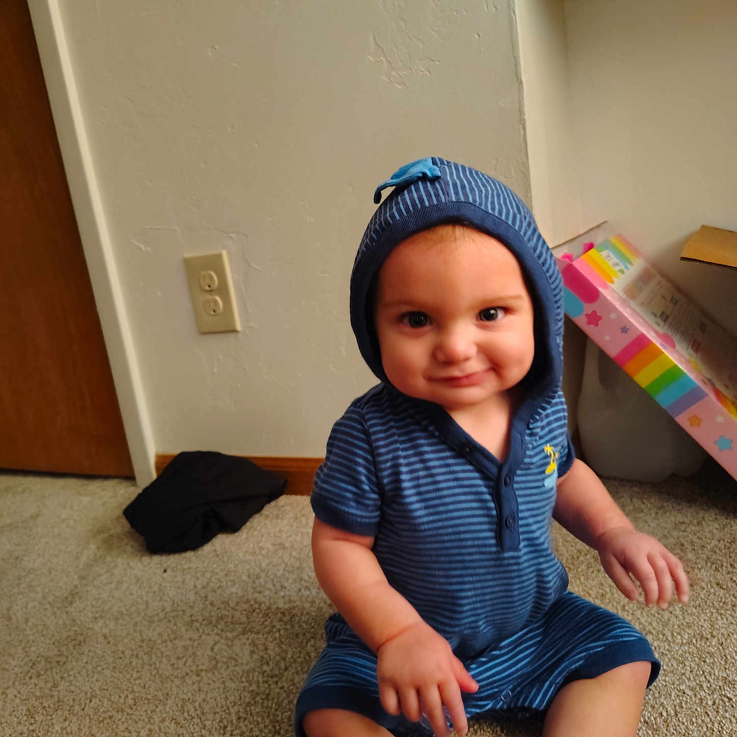 Luther joined the competition — help win amazing prizes! baby, child, toddler, sitting, smiling, happy, portrait, face, blue_outfit, striped_clothing, hat, hood, carpet, indoor, home, door, wall, electrical_outlet, colorful_package, bare_feet