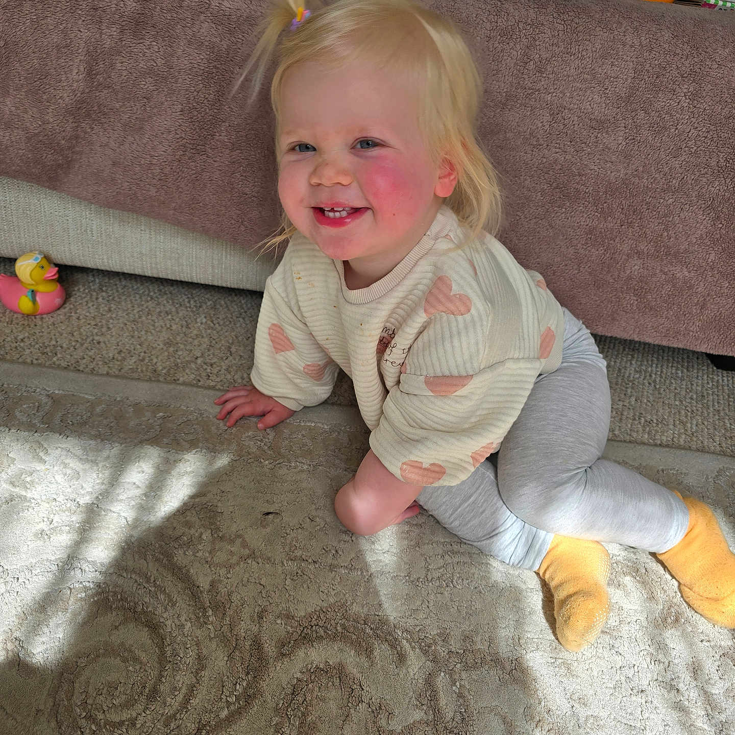 Evie Joy is registered to the contest to win money with this photo: blonde_hair, carpet, child, cozy, crawling, face, furniture, happy, hearts, indoor, living_room, person, playful, plush, pony_tail, smiling, sweater, toddler, toy_duck, yellow_socks