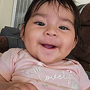 Zoe is registered to the contest to win money with this photo: baby, child, smile, face, eyes, hand, onesie, clothing, indoors, couch, portrait, close_up, happy, adorable, cheeks, hair, mouth, toddler, sitting, home