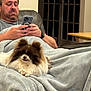 man, dog, blanket, couch, phone, indoor, relaxed, brown, white, fluffy, furniture, table, watch, casual, sitting, technology, comfort, pet, home, leather