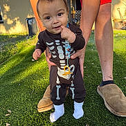 Josiah joined the competition — help win amazing prizes! baby, child, skeleton_onesie, grass, outdoor, sunlight, person, adult_hands, feet, socks, curious, standing, supporting_hands, shorts, slippers, lawn, daylight, cute, young_child, portrait