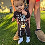 baby, child, skeleton_onesie, grass, outdoor, sunlight, person, adult_hands, feet, socks, curious, standing, supporting_hands, shorts, slippers, lawn, daylight, cute, young_child, portrait