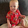 toddler, child, smiling, sitting, red_clothing, floral_pattern, blond_hair, barefoot, couch, blanket, indoor, happy, cute, portrait, person, baby, casual, relaxed, young_child, home