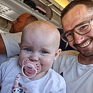 Kelyana participe au concours pour gagner de l'argent avec cette photo : baby, pacifier, man, glasses, airplane, passenger, seat, overhead_compartment, luggage, smile, child, white_shirt, beads, earring, portrait, indoor, travel, family, happy, people