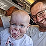 baby, pacifier, man, glasses, airplane, passenger, seat, overhead_compartment, luggage, smile, child, white_shirt, beads, earring, portrait, indoor, travel, family, happy, people