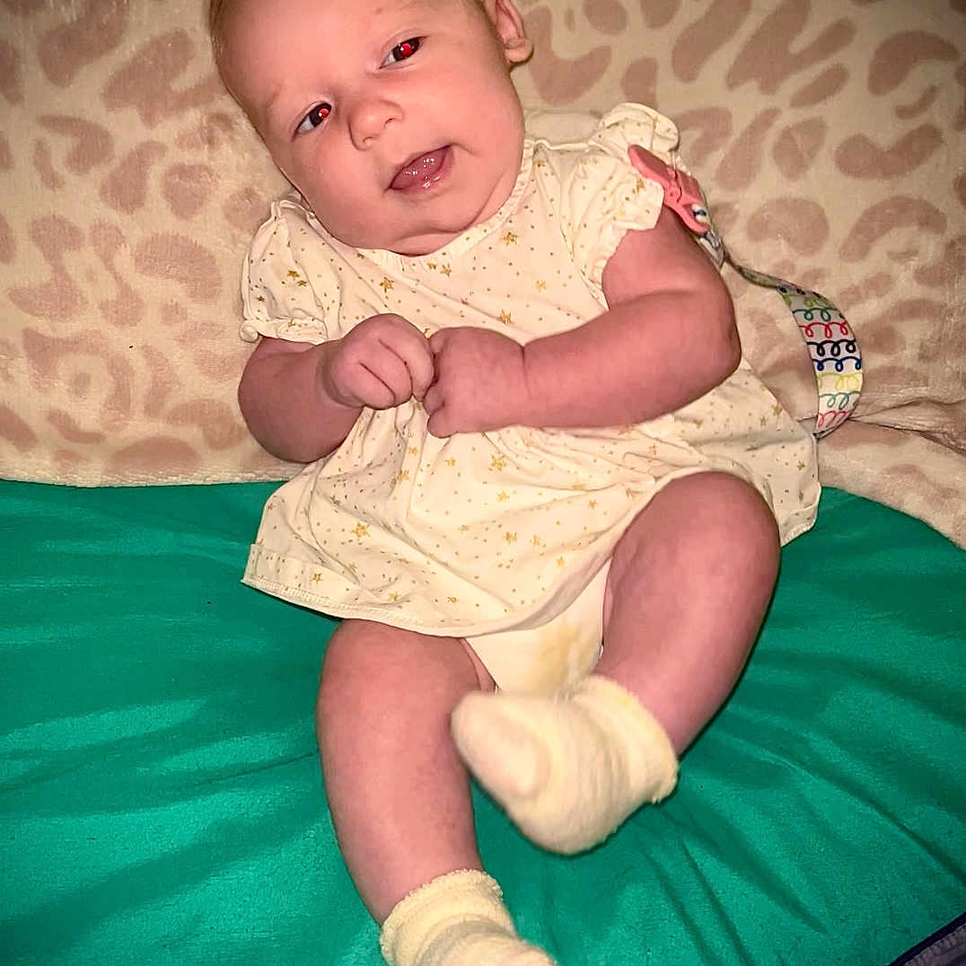 Brooklyn is registered to the contest to win money with this photo: baby, blanket, child, clothing, cute, expression, face, hands, indoors, infant, pattern, person, portrait, sitting, skin, smile, socks, teal_surface, yellow_dress, young_child