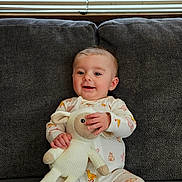 Ginny joined the competition — help win amazing prizes! baby, child, pajamas, stuffed_animal, couch, gray_couch, smiling, happy, indoor, window_blinds, knitted_toy, feet, sitting, infant, cute, relaxed, home, soft_toy, childrens_clothing, cozy