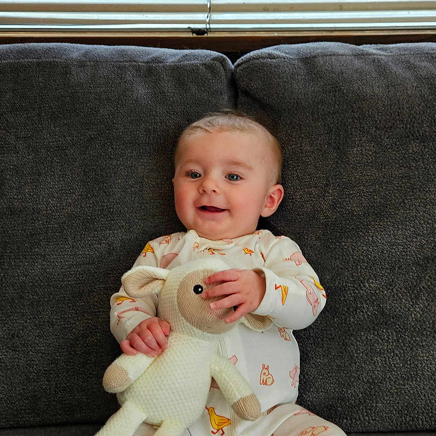 Ginny joined the competition — help win amazing prizes! baby, child, childrens_clothing, couch, cozy, cute, feet, gray_couch, happy, home, indoor, infant, knitted_toy, pajamas, relaxed, sitting, smiling, soft_toy, stuffed_animal, window_blinds