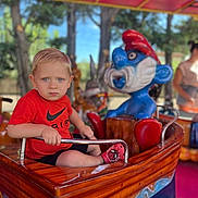 Gianni participe au concours pour gagner de l'argent avec cette photo : toddler, child, amusement_ride, boat, red_shirt, nike_logo, blue_eyes, shorts, spiderman_socks, metal_bar, wooden_surface, outdoor, trees, daylight, ride_character, person, background_people, playground, fun, park