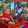 toddler, child, amusement_ride, boat, red_shirt, nike_logo, blue_eyes, shorts, spiderman_socks, metal_bar, wooden_surface, outdoor, trees, daylight, ride_character, person, background_people, playground, fun, park