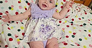 Ariel is registered to the contest to win money with this photo: baby, infant, girl, smiling, headband, bow, floral_dress, blanket, polka_dots, bed, wooden_bed, happy, arms_outstretched, milestone, text_overlay, indoor, child, cute, lying_down, portrait