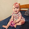 baby, child, smiling, toy, pink, headband, flower, dress, plaid, bed, bedding, pillow, indoor, person, happy, sitting, cute, infant, playful, blue