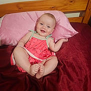 Ariel joined the competition — help win amazing prizes! baby, child, infant, smiling, pink_pillow, bed, bedding, red_blanket, wooden_headboard, cute_outfit, watermelon_dress, happy, sitting, indoors, person, portrait, skin, face, feet, hand