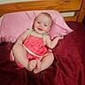 baby, child, infant, smiling, pink_pillow, bed, bedding, red_blanket, wooden_headboard, cute_outfit, watermelon_dress, happy, sitting, indoors, person, portrait, skin, face, feet, hand