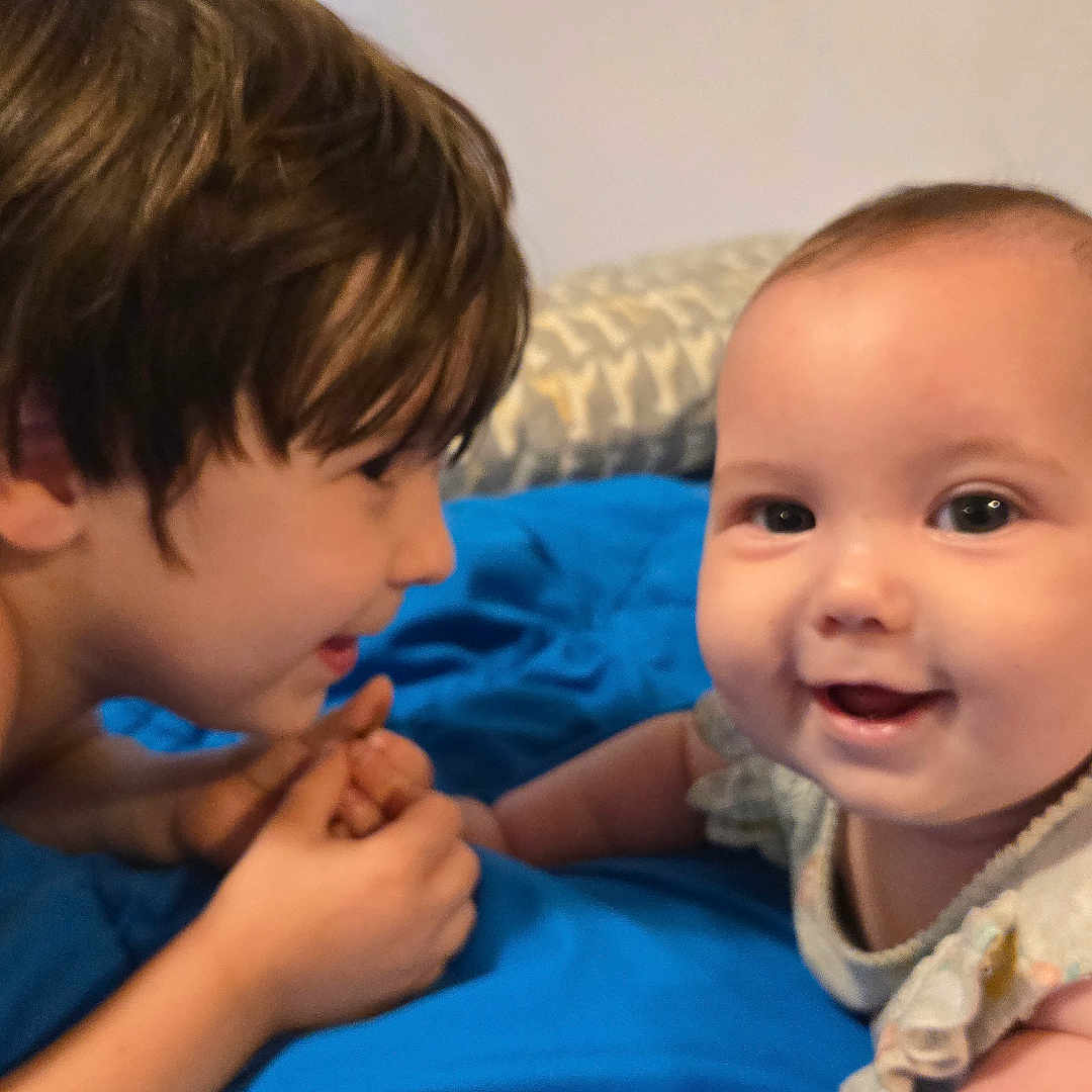 Arabella is registered to the contest to win money with this photo: baby, baby_clothes, bedroom, blue_blanket, child, clothing, curly_hair, cute, face, family, happy, indoor, infant, interaction, person, portrait, sibling, skin, smile, toddler