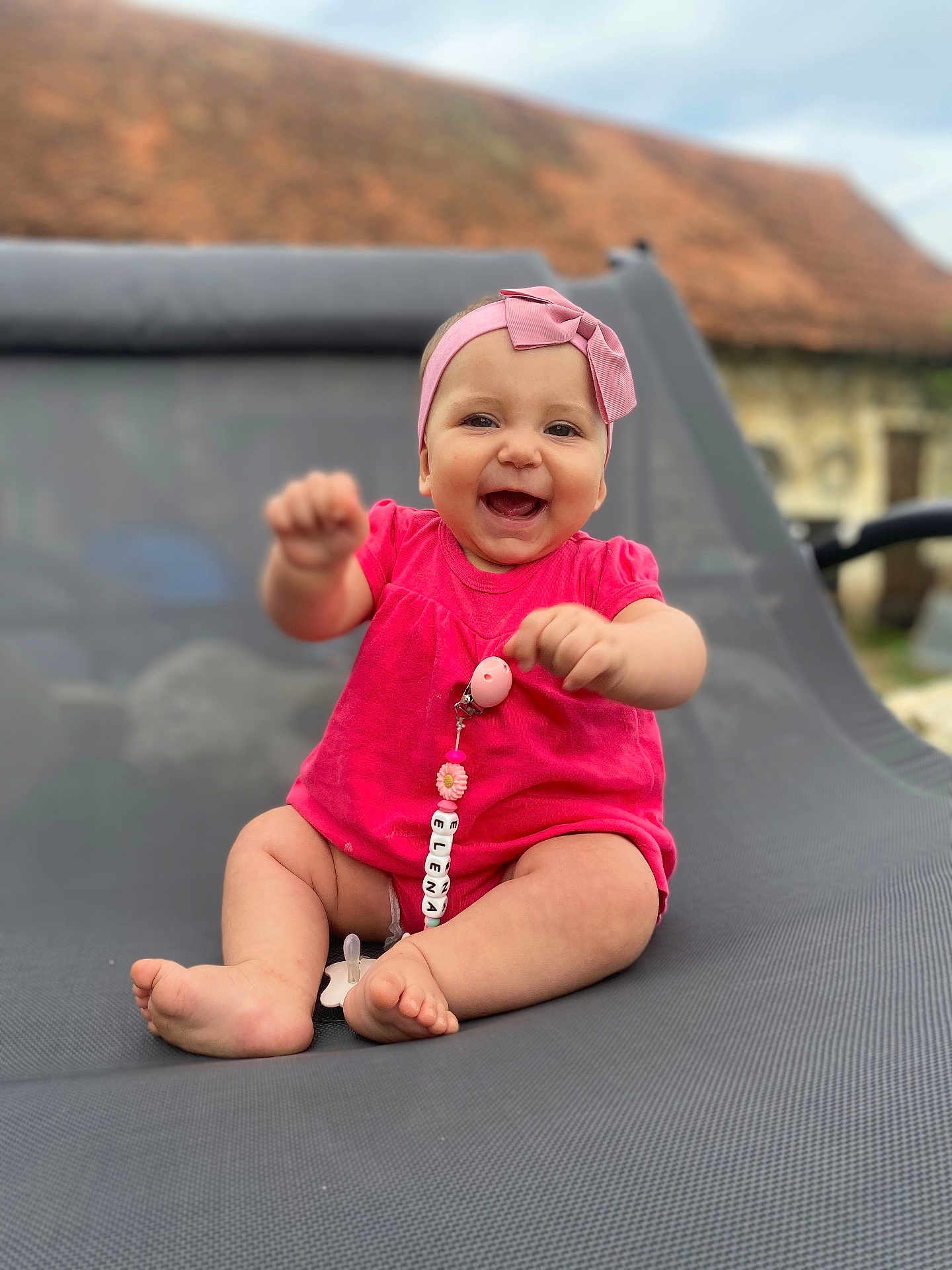 Elena participe au concours pour gagner de l'argent avec cette photo : baby, child, smiling, pink_clothing, headband, pacifier, outdoor, lounge_chair, happy, infant, cute, person, sitting, toy, blurred_background, skin, face, legs, feet, daylight